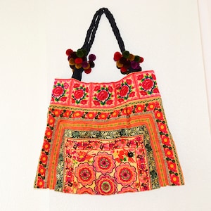 May include: A colorful embroidered tote bag with a pink, red, and green floral pattern. The bag has black straps with colorful pom-poms.