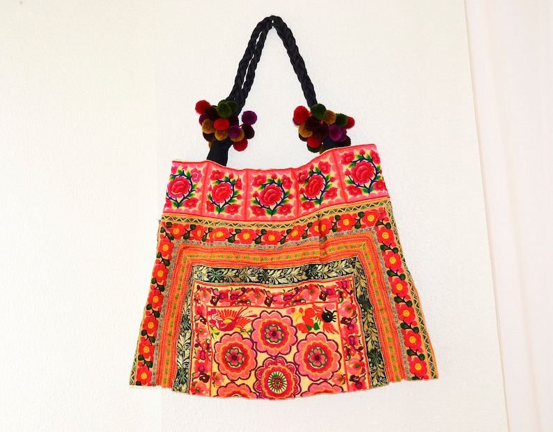 May include: A colorful embroidered tote bag with a pink, red, and green floral pattern. The bag has black straps with colorful pom-poms.