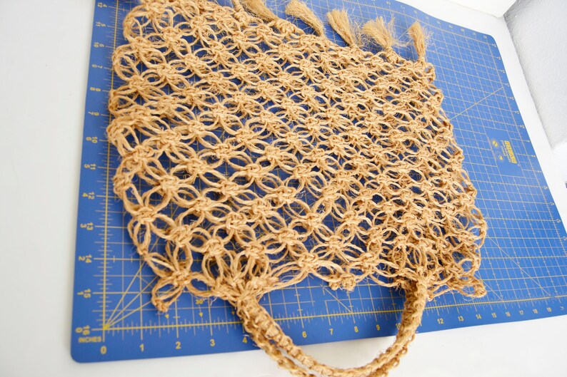 May include: A tan crocheted tote bag with long fringe tassels. The bag is made of natural fibers and has a woven texture.
