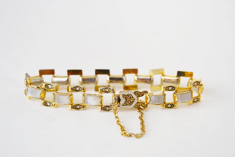 May include: A gold-tone bracelet with a white enamel design. The bracelet has a decorative clasp and a chain.