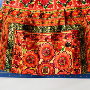 May include: A close-up of a colorful embroidered fabric with a floral pattern. The fabric is a mix of red, orange, yellow, green, and black. The embroidery is intricate and detailed.