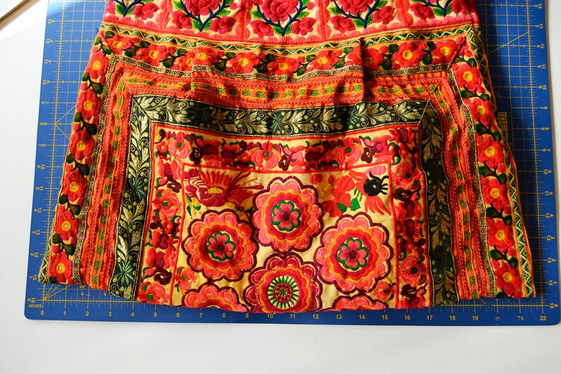 May include: A close-up of a colorful embroidered fabric with a floral pattern. The fabric is a mix of red, orange, yellow, green, and black. The embroidery is intricate and detailed.