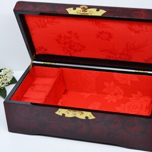 Peut inclure: Une bo&icirc;te &agrave; bijoux rectangulaire ouverte avec une finition rouge fonc&eacute; brillante. L'int&eacute;rieur est doubl&eacute; de tissu rouge avec un motif floral. La bo&icirc;te est orn&eacute;e de ferrures dor&eacute;es et est con&ccedil;ue pour ranger des bijoux.