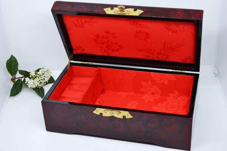 Peut inclure: Une bo&icirc;te &agrave; bijoux rectangulaire ouverte avec une finition rouge fonc&eacute; brillante. L'int&eacute;rieur est doubl&eacute; de tissu rouge avec un motif floral. La bo&icirc;te est orn&eacute;e de ferrures dor&eacute;es et est con&ccedil;ue pour ranger des bijoux.