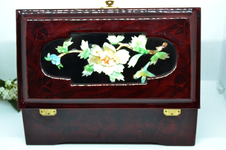 Peut inclure: Une bo&icirc;te &agrave; bijoux rectangulaire en bois laqu&eacute;, de couleur bordeaux fonc&eacute;. La bo&icirc;te pr&eacute;sente un panneau d&eacute;coratif central avec un motif floral en nacre et un fermoir en laiton.