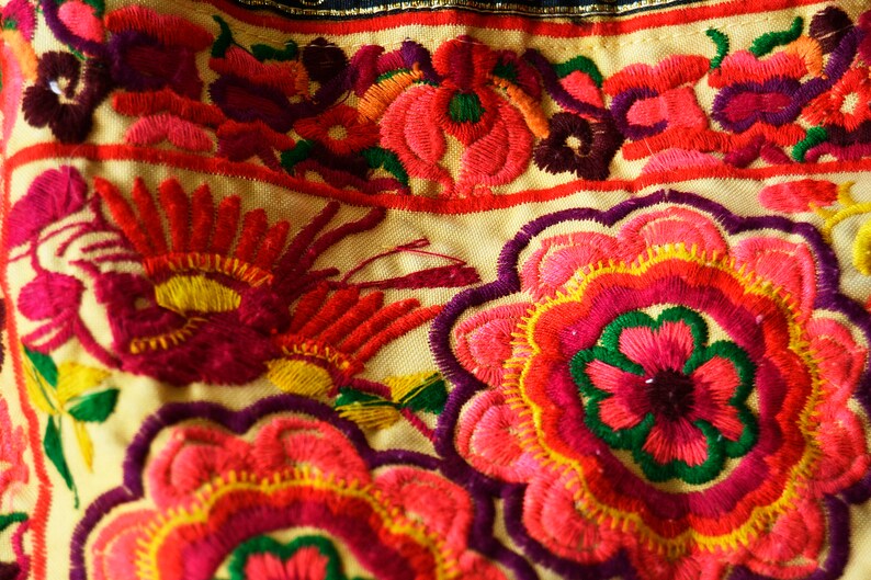 May include: Close-up of colorful embroidered fabric with a floral pattern. The fabric is cream-colored with red, pink, yellow, green, and purple embroidery.
