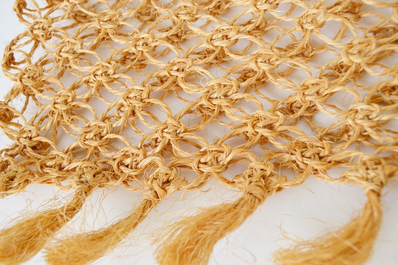 May include: A close-up of a beige macrame wall hanging with fringe tassels. The macrame is made with a natural fiber rope and has a diamond pattern.