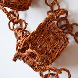 May include: A brown woven chain with two small woven baskets attached. The chain is made of intertwined strands of brown material, forming a series of connected loops. The baskets are woven in a similar style, with a slightly more open weave.