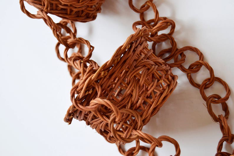 May include: A brown woven chain with two small woven baskets attached. The chain is made of intertwined strands of brown material, forming a series of connected loops. The baskets are woven in a similar style, with a slightly more open weave.