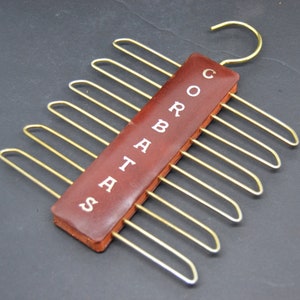 May include: A brown leather tie rack with gold metal hooks. The rack has the word "CORBATAS" written on it.