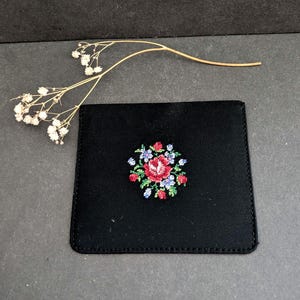 Op de afbeelding: Een zwarte vierkante portemonnee met een bloemenkruissteekontwerp in rood, roze, blauw en groen. De portemonnee is gemaakt van een glad, donker materiaal en wordt op een donkere ondergrond getoond.
