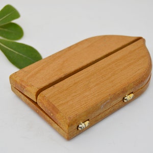 May include: A small wooden box with a hinged lid and a decorative arch shape. The box is made of light brown wood and has two small gold hinges.