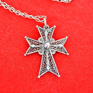 May include: A silver filigree cross pendant on a silver chain. The pendant is a detailed design with a small round bead in the center. The cross is hanging on a red background.