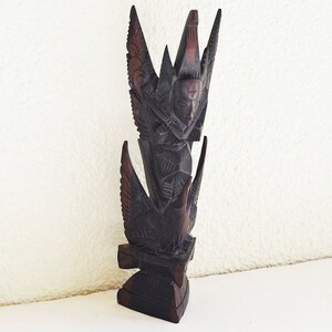May include: A dark brown wooden sculpture of a mythical creature with wings and a human-like face. The sculpture is carved in a detailed and intricate style.