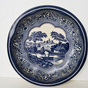 Puede incluir: Un plato de cerámica azul y blanco con un borde floral y una escena de un río con un puente y una casa en el fondo.
