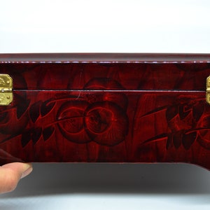 Peut inclure: Une bo&icirc;te rectangulaire en bois laqu&eacute; avec une finition rouge fonc&eacute; et des motifs floraux d&eacute;coratifs. La bo&icirc;te comporte deux fermoirs et charni&egrave;res dor&eacute;s orn&eacute;s. La bo&icirc;te est tenue par une main.