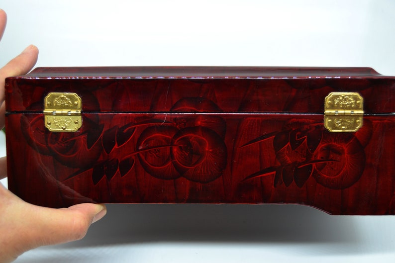 Peut inclure: Une bo&icirc;te rectangulaire en bois laqu&eacute; avec une finition rouge fonc&eacute; et des motifs floraux d&eacute;coratifs. La bo&icirc;te comporte deux fermoirs et charni&egrave;res dor&eacute;s orn&eacute;s. La bo&icirc;te est tenue par une main.