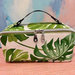 May include: A rectangular toiletry bag featuring a white base with a green monstera leaf pattern. The bag includes a dark brown top handle, a silver-toned zipper closure with two metal pulls, and a structured shape for storing personal items.