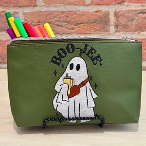 May include: An olive green zippered pouch featuring an embroidered white ghost character holding a yellow tumbler and wearing a brown crossbody bag. The text BOO-JEE is embroidered above the ghost, and several colorful markers are tucked inside.