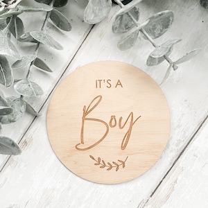 Puede incluir: Un letrero redondo de madera con el texto "It's a Boy" en letra cursiva. El letrero está decorado con una pequeña rama de hojas.
