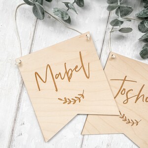 Puede incluir: Dos letreros de madera con los nombres "Mabel" y "Josh" grabados en ellos. Cada letrero tiene un peque&ntilde;o dise&ntilde;o de rama con hojas debajo del nombre. Los letreros est&aacute;n colgados de una cuerda.