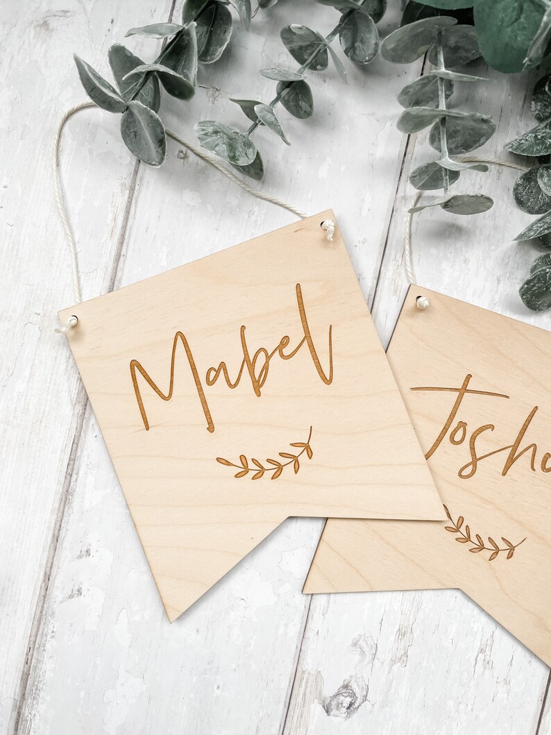 Puede incluir: Dos letreros de madera con los nombres "Mabel" y "Josh" grabados en ellos. Cada letrero tiene un peque&ntilde;o dise&ntilde;o de rama con hojas debajo del nombre. Los letreros est&aacute;n colgados de una cuerda.
