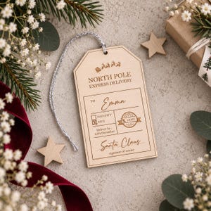 May include: A rectangular wooden gift tag with a silver string, featuring the engraved text NORTH POLE EXPRESS DELIVERY, TO: Emma, a checked NICE box, a circular stamp with 12002, Deliver by: 25th December, and the signature Santa Claus.