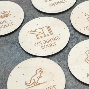 Puede incluir: Un conjunto de ocho etiquetas redondas de madera con varias ilustraciones y texto. Las etiquetas presentan im&aacute;genes de un coche, un dinosaurio, un elefante, un libro para colorear, y m&aacute;s. El texto en las etiquetas incluye "Animales", "Coches y camiones", "Libros para colorear", y "Dinosaurios".