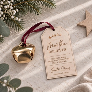 May include: A gold-colored metal jingle bell attached by a dark red ribbon to a rectangular wooden tag. The tag features an engraved silhouette of a sleigh with reindeer, the name Martha, the text BELIEVES, and the phrase THIS BELL STILL RINGS FOR THOSE WHO TRULY BELIEVE, followed by Santa Claus and signature of sender.
