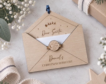 Könnte beinhalten: Ein Briefumschlag-Anhänger aus hellem Holz mit der Gravur Dear Santa, Daniel's CHRISTMAS WISH LIST und einer Silhouette eines Schlittens mit Rentieren. Ein kreisförmiges Holzsiegel mit der Aufschrift N. 12003 verschließt die Klappe mit einer dunkelblauen Schleife.