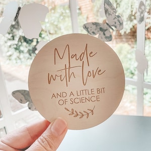 Puede incluir: Un letrero redondo de madera con el texto "Made with love and a little bit of science" en una fuente marrón. El letrero tiene un pequeño diseño de hojas en la parte inferior.