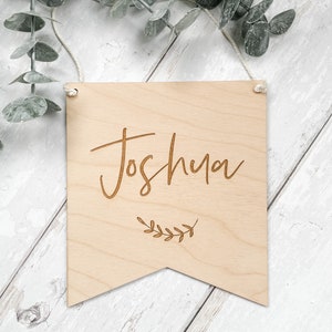 Puede incluir: Un letrero de madera con el nombre "Joshua" grabado en letra cursiva. El letrero tiene forma de bander&iacute;n y tiene un peque&ntilde;o dise&ntilde;o de hoja debajo del nombre.