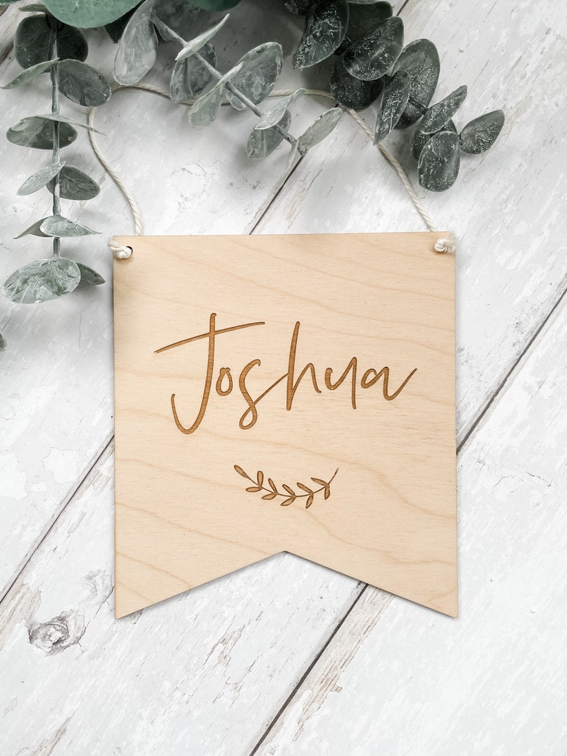Puede incluir: Un letrero de madera con el nombre "Joshua" grabado en letra cursiva. El letrero tiene forma de bander&iacute;n y tiene un peque&ntilde;o dise&ntilde;o de hoja debajo del nombre.