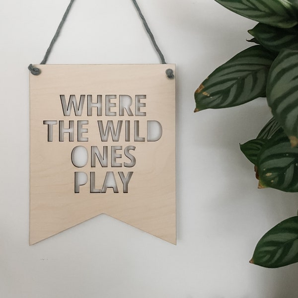 May include: A wooden sign with the text "Where the Wild Ones Play" cut out in the shape of the letters. The sign is hanging from a gray string and is against a white wall.