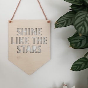 May include: Wooden sign with the text "Shine Like The Stars" cut out in a white font. The sign is hanging from a string and is in front of a white wall.