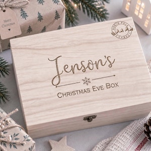 Puede incluir: Una caja rectangular de madera clara con tapa abatible y cierre metálico. La tapa presenta el texto grabado Jenson's Christmas Eve Box, un pequeño icono de copo de nieve y un sello circular de North Pole Express con un reno.