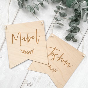 Puede incluir: Dos letreros de madera con los nombres "Mabel" y "Joshua" grabados en ellos. Cada letrero tiene una peque&ntilde;a rama de hojas en la parte inferior.