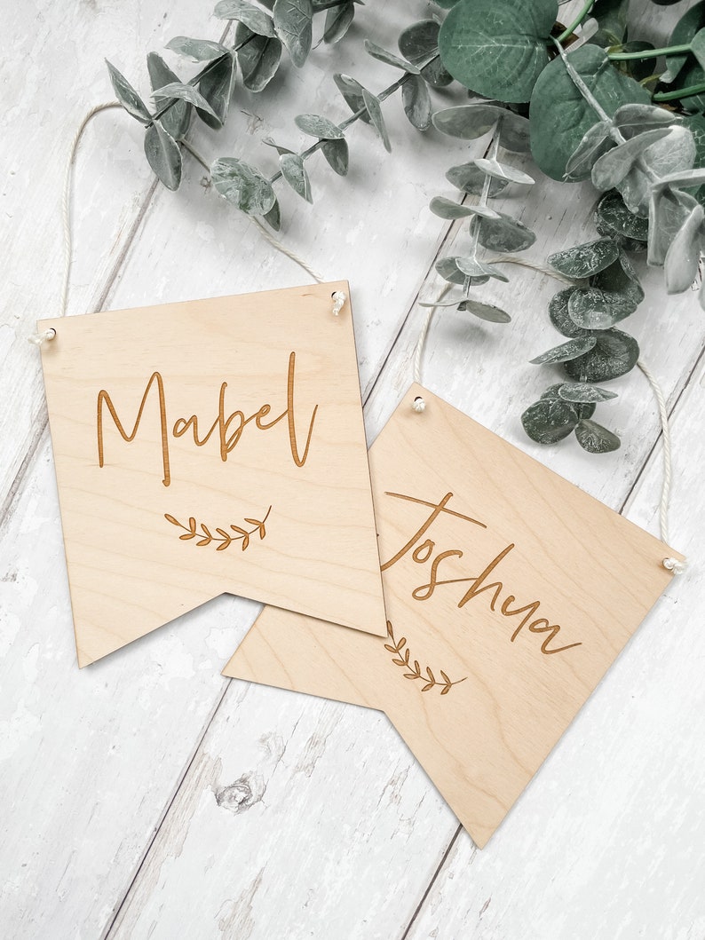 Puede incluir: Dos letreros de madera con los nombres "Mabel" y "Joshua" grabados en ellos. Cada letrero tiene una peque&ntilde;a rama de hojas en la parte inferior.