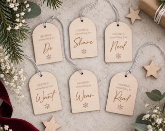 Op de afbeelding: Zes boogvormige cadeaulabels van licht hout met zilverkleurig touw, elk gegraveerd met 'GEORGE, something to' gevolgd door 'Do', 'Share', 'Need', 'Want', 'Wear' of 'Read', en een klein sneeuwvlokpictogram op een neutrale ondergrond.