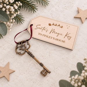Puede incluir: Una llave antigua de color bronce unida por una cinta roja oscura a una etiqueta rectangular de madera. La etiqueta presenta el texto grabado "Santa's Magic Key TO FINLEY'S HOUSE" junto a un gráfico de trineo y renos.