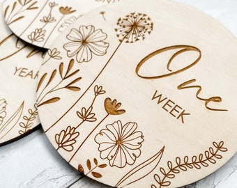 May include: Three wooden baby milestone discs with a floral design. The discs read "Seven Months", "One Year", and "One Week".