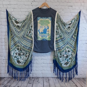 May include: A dark grey t-shirt with a vintage concert poster design, paired with a patterned shawl featuring blue, green, and cream floral and geometric designs. The shawl has blue fringe.