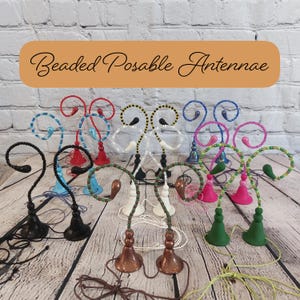 May include: A collection of beaded, posable antennae in various colors, including black, red, white, blue, pink, and green. Each antenna features a decorative base and a curved, beaded top. The text "Beaded Posable Antennae" is displayed in an orange banner.
