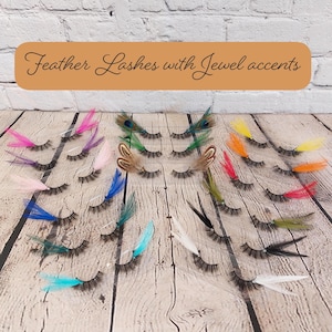 Può includere: Una collezione di ciglia finte con accenti di piume in vari colori, tra cui rosa, blu, verde e giallo. Il testo "Feather Lashes with Jewel accents" è visualizzato su uno striscione marrone sopra le ciglia. Le ciglia sono disposte su una superficie di legno.