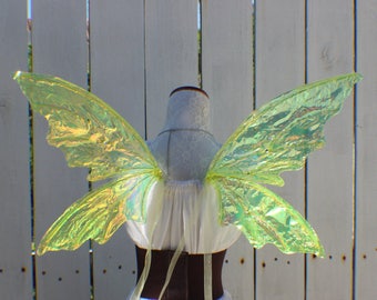 May include: A pair of iridescent green fairy wings with a shimmery finish. The wings are attached to a white bodice with a brown belt.