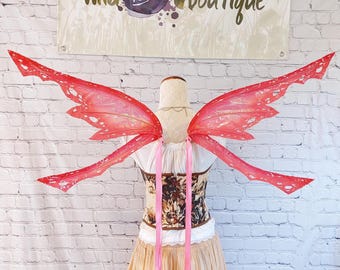 May include: Large, translucent red fairy wings with jagged, insect-like edges and gold vein detailing. The wings are attached to a white bodice featuring a dark floral pattern and a long, flowing beige skirt, with two long pink ribbons hanging down.