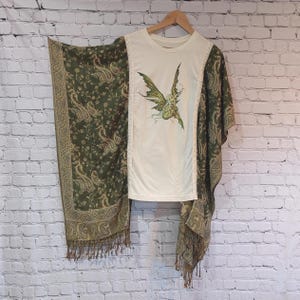 May include: A white t-shirt with a fairy graphic, paired with a green and gold paisley shawl. The shawl has a fringe trim. The shirt is hanging on a wooden hanger against a white brick wall.