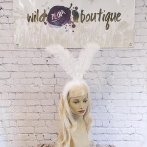 May include: A white headband featuring two tall, upright ostrich feathers arranged to resemble rabbit ears. The headband is worn on a mannequin head with long, wavy blonde hair, positioned in front of a sign that reads wild plum boutique.