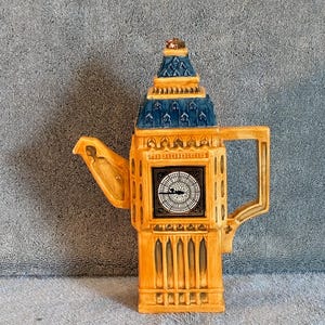 May include: A ceramic teapot shaped like the Elizabeth Tower, commonly known as Big Ben. The structure features a tan body with architectural details, a blue roof, and a clock face on the front. It includes a handle on the right and a spout on the left.