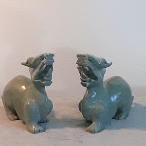 May include: Two green jade stone sculptures of mythical creatures with open mouths and sharp teeth. The sculptures are facing each other and are in a seated position.
